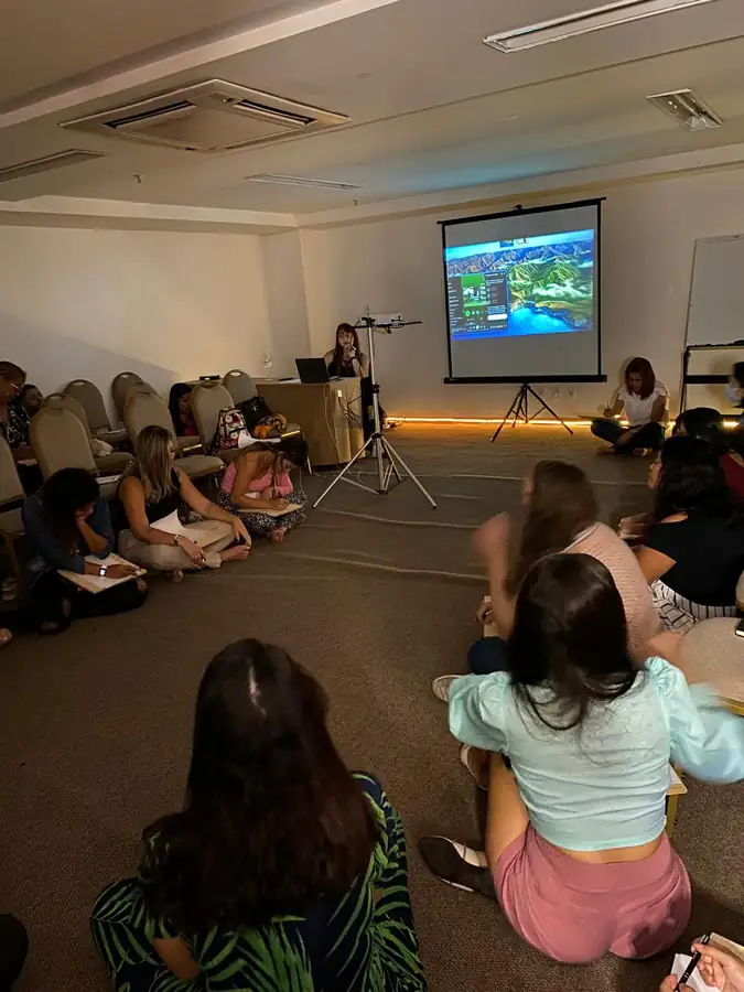 Participantes sentados no chão em sala de treinamento, com Ana Paula conduzindo a atividade ao lado do telão